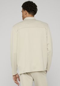 Veste beige clair à manches longues et coupe droite. Elle présente une couture de yoke au dos et des surpiqûres discrètes pour une texture lisse.