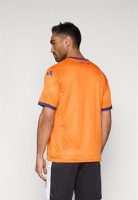 Tricou sport portocaliu cu accente mov pe mâneci, textură plasă și textul „FIORENTINA” pe spate. Purtat cu pantaloni scurți negri.