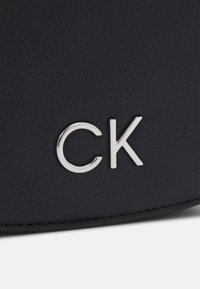 Czarna teksturowana skóra z metalicznym srebrnym detalem w postaci logo "CK", podkreślająca gładkie i wyrafinowane wykończenie powierzchni.