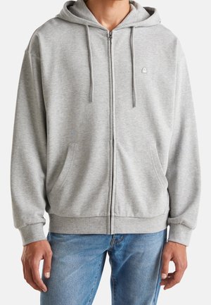 Mand iført en lysegrå lynlåshættetrøje med forlommer og snore, kombineret med blå jeans, vist fra skuldre til hofter.