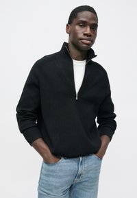 Μαύρο πλεκτό ζακέτα με φερμουάρ και ψηλό γιακά· ριμπ μανσέτες και τελείωμα. Φορεμένο πάνω από λευκό μπλουζάκι. Συνδυασμένο με ξεθωριασμένο μπλε τζιν.