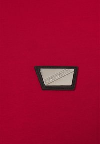 Un tissu rouge à texture striée présente un badge triangulaire noir avec un accent argenté, inscrit avec "ANTONY MORATO."