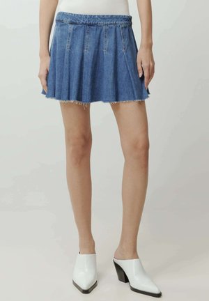 Vrouw met een korte blauwe denim plooirok met onafgewerkte zoom en witte muiltjes met hak, staand tegen een neutrale achtergrond.