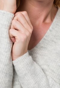 Pull gris clair en tricot texturé, avec des manches longues, des poignets côtelés et un design en V, les mains croisées près du col.