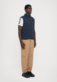 Navy puffer vest with a high collar and two side pockets, worn over a white shirt. Paired with beige cargo pants and black sneakers.