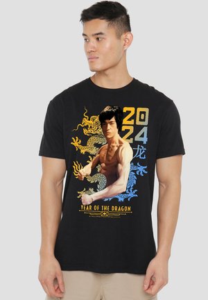 Man wearing black t-shirt with graphic of a shirtless martial artist, yellow and blue dragon, and text "2024 Year of the Dragon."