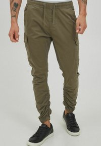 Oliwkowe spodnie cargo wykonane z lekkiego materiału, z elastycznym pasem, sznurkiem, bocznymi kieszeniami i ściągaczami na mankietach. Czarne sneakersy.