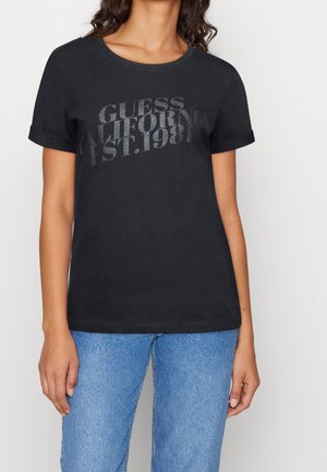 Femme portant un t-shirt noir aux manches retroussées avec un texte délavé "GUESS CALIFORNIA EST. 1981" associé à un jean bleu.