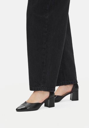 Czarne skórzane buty na klockowym obcasie z zaostrzonym noskiem i błyszczącym elementem na czubku, zestawione z ciemnymi, szerokimi spodniami typu jeans.