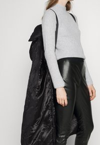Grauer gerippter Pullover mit hohem Kragen, kombiniert mit schwarzen Lederhosen; ein schwarzer gepolsterter Mantel hängt über dem Arm.