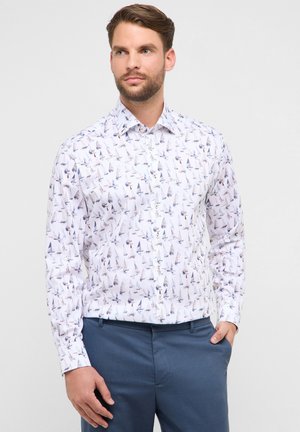 Uomo con capelli castani e barba, che indossa una camicia bianca a maniche lunghe con una piccola stampa di barche a vela e pantaloni blu, in piedi davanti a uno sfondo chiaro.