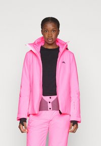 Chaqueta aislante de color rosa brillante con un alto cuello, cremallera frontal y cordón. Logo negro en el pecho. Acompañada de pantalones rosas a juego.