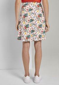 Femme portant une jupe A-line fleurie avec des motifs rouges, jaunes et verts, associée à des baskets blanches, se tenant contre un fond gris uni.