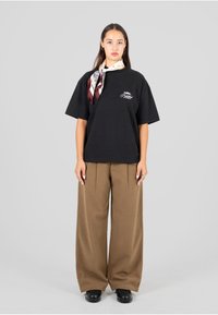 Czarna, oversize koszulka z logo, noszona z wzorzystą jedwabną chustą. W połączeniu z szerokimi brązowymi spodniami i czarnymi butami na platformie.