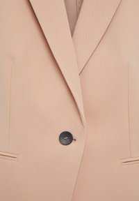 Světle béžový blazer s klopovým límcem, zapínáním na jeden černý knoflík a dvěma předními kapsami. Hladká tkanina s přiléhavým střihem.