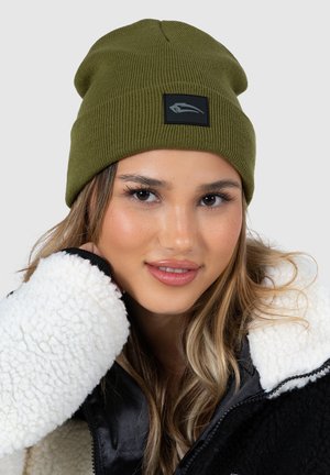 Oliven grøn, ribbet strikket beanie med en sort firkantet applikation med logo. Modellen bærer en hvid sherpa-futteret jakke med sorte accenter.