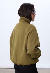 Olivgrüne, kragenlose Jacke mit lockerer Passform, die einen gerafften Saum und umschlagbare Ärmel hat. Glattes Material und minimalistisches Design.