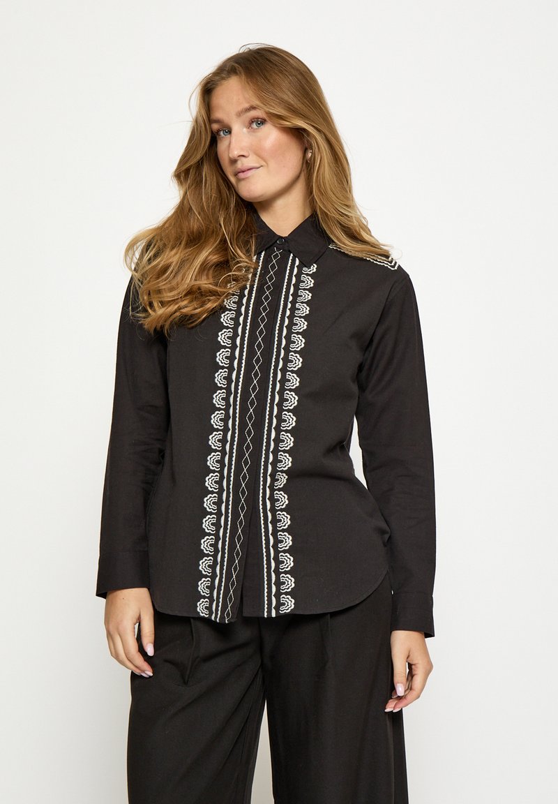 Čierna košeľa s dlhými rukávmi s bielymi vyšívanými vzormi vpredu a na golieri, s zakriveným lemom a zapínaním na gombíky.