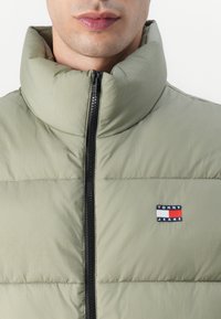 Padded vest in light green, featuring a high collar and black zipper. Includes a blue, white, and red embroidered "TOMMY JEANS" logo.