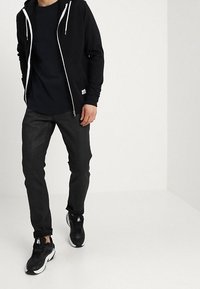 Černá zipová mikina s bílými šnůrkami, nošená přes tmavé tričko. Slim-fit černé džíny a černé tenisky s bílými akcenty doplňují vzhled.