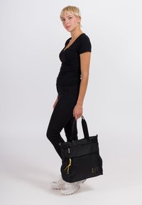 Zwarte tas met twee handvatten aan de bovenkant, textuurstof, ritszakken, gele accenten en een prominent weergegeven logo. Gecombineerd met een zwarte outfit.