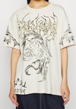 Vrouw draagt een oversized off-white T-shirt met abstracte zwart-gouden grafische ontwerpen en onleesbare tekst, gecombineerd met een donkere spijkerrok.