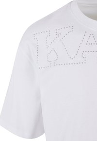 Balta kokvilnas T-kreklu ar teksturētu sudraba kniedžu rakstu burtu formā priekšpusē. Īsas piedurknes ar noapaļotu kakla izgriezumu.