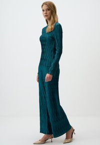 Salopette lunga teal a maniche lunghe con motivi verticali, corpetto aderente e pantaloni svasati. Tessuto liscio e lucido. Indossata con tacchi di colore chiaro.