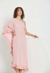 Mujer de cabello rizado que lleva un vestido rosa de un solo hombro con bordes con volantes, de pie con una mano en la cadera y la otra sobre un accesorio blanco redondeado.