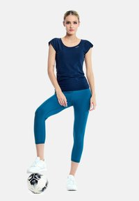 Tricou sportiv cu mânecă scurtă de culoare bleumarin, asociat cu colanți tec cu lungime până la genunchi. Modelul stă pe o minge de fotbal, purtând adidași albi.