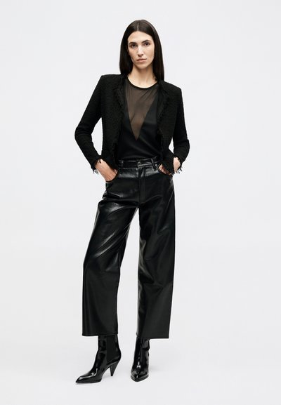 Žena v čiernom textúrovanom blejzri, priehľadnom čiernom topu, kožených širokých nohaviciach s vysokým pásom a špicatých čiernych členkových topánkach stojí s rukami v vreckách.