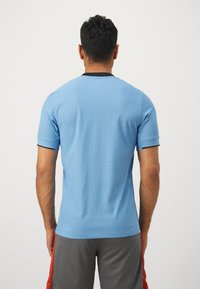 Maglietta atletica a maniche corte di colore blu chiaro con motivo testurizzato, colletto a girocollo e dettagli neri sulle maniche. Indossata con pantaloni corti grigi.