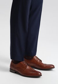Braune Leder-Dressschuhe mit dekorativen Perforationen, flacher schwarzer Sohle, Schnürsenkeln und kombiniert mit marineblauen gemusterten Hosen.
