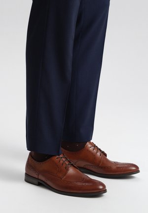 Brown leather dress shoes with decorative perforations, flat black sole, laces, and paired with navy patterned trousers.
