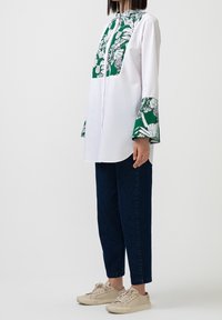 Camicia bianca con stampa floreale verde sul davanti e sui polsini, dotata di un collo alto e maniche lunghe. Abbinata a jeans blu scuri e sneakers beige.
