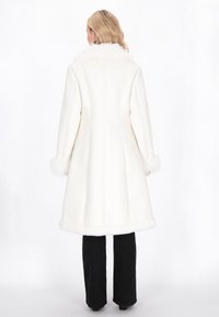White long coat with a faux fur collar and cuffs, featuring a structured design, clean lines, and a smooth texture. Worn over black pants.