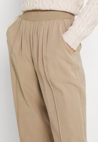 Béžové kalhoty s elastickým pasem, s jemnými záhyby a bočními kapsami. Hladká textura s uvolněným, rovně střiženým střihom.