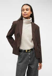 Brūns žakets, izgatavots no strukturēta auduma, valkāts virs gaiši pelēka augstā apkakle, apvienots ar melniem džinsiem ar augstu jostasvietu un melnu jostu ar zelta sprādzi.