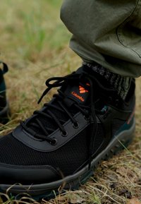 Czarne sneakersy do użytku na zewnątrz z siateczkowym i syntetycznym wykończeniem, z pomarańczowymi akcentami i czarnymi sznurowadłami, umieszczone na trawiastej powierzchni.