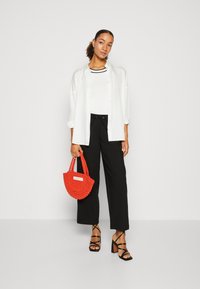 Hvit oversized skjorte med knapper over en tettsittende hvit topp, paret med sorte bukser og sorte sandaler med stropper. Holder en liten rød vevd veske.