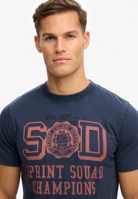 Camiseta de color azul marino con texto gráfico rojo "SOD SPRINT SQUAD CHAMPIONS" y un emblema circular, confeccionada en suave material de algodón.