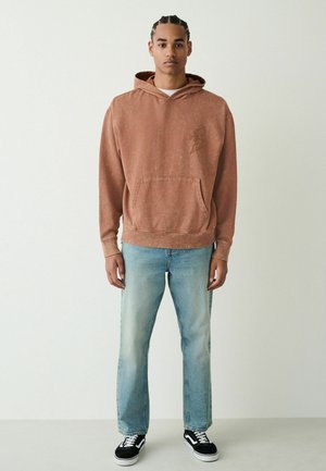 Junger Mann, der einen verblassten braunen Hoodie, hellblaue Jeans und schwarz-weiße Turnschuhe trägt und vor einer schlichten weißen Wand steht.