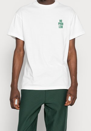 Homme portant un t-shirt blanc à manches courtes avec le texte vert "NO PROBLEM" en haut à gauche et un pantalon vert foncé, debout devant un fond uni.