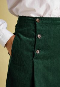 Grüner Cordrock mit vertikalen Rippungen, einer Seitentasche und drei braunen Knopfapplikationen an der Vorderseite.