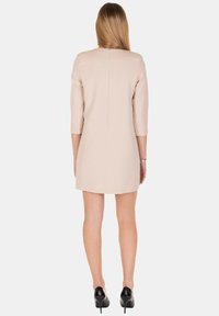 Vestito corto beige con maniche a tre quarti e taglio dritto. Presenta una cucitura posteriore, abbinato a scarpe nere con tacco alto.
