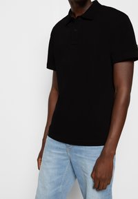 Czarna koszulka typu polo wykonana z teksturowanej tkaniny, z kołnierzykiem, listwą z trzema guzikami i krótkimi rękawami, zestawiona z jasnymi dżinsami.