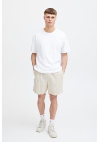 Tricou alb din bumbac cu mâneci scurte, asortat cu pantaloni scurți bej texturați și adidași albi, pe un fundal simplu.