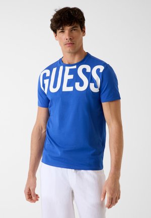 Homme portant un t-shirt bleu avec un grand texte blanc « GUESS » sur la poitrine et un pantalon blanc, debout devant un fond uni.