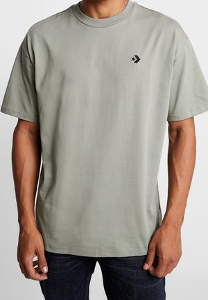 Mężczyzna ubrany w jasnoszary t-shirt z krótkim rękawem z małym czarnym logo Converse na lewej piersi oraz ciemne jeansy.