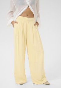 Pantaloni larghi a plissettatura gialla con vita alta e tessuto liscio, abbinati a una camicia bianca trasparente con bottoni.
